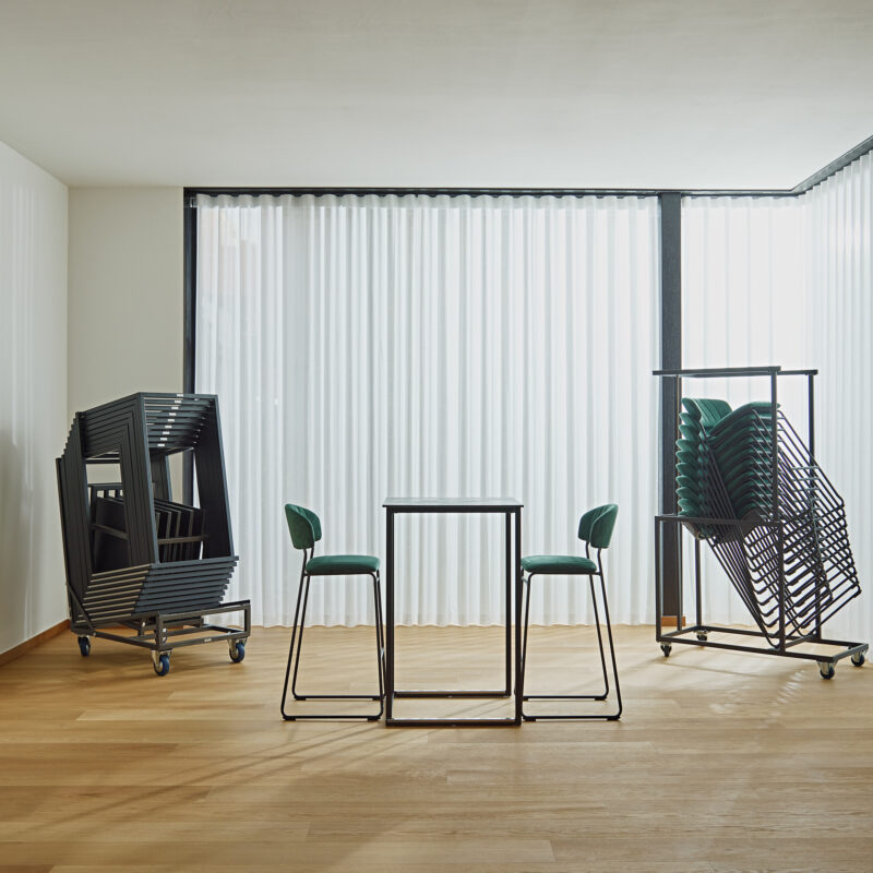 Chariot de transport avec tables hautes Kubo empilées et tabourets de bar Teddy, avec une table haute et deux tabourets de bar disposés devant le chariot.